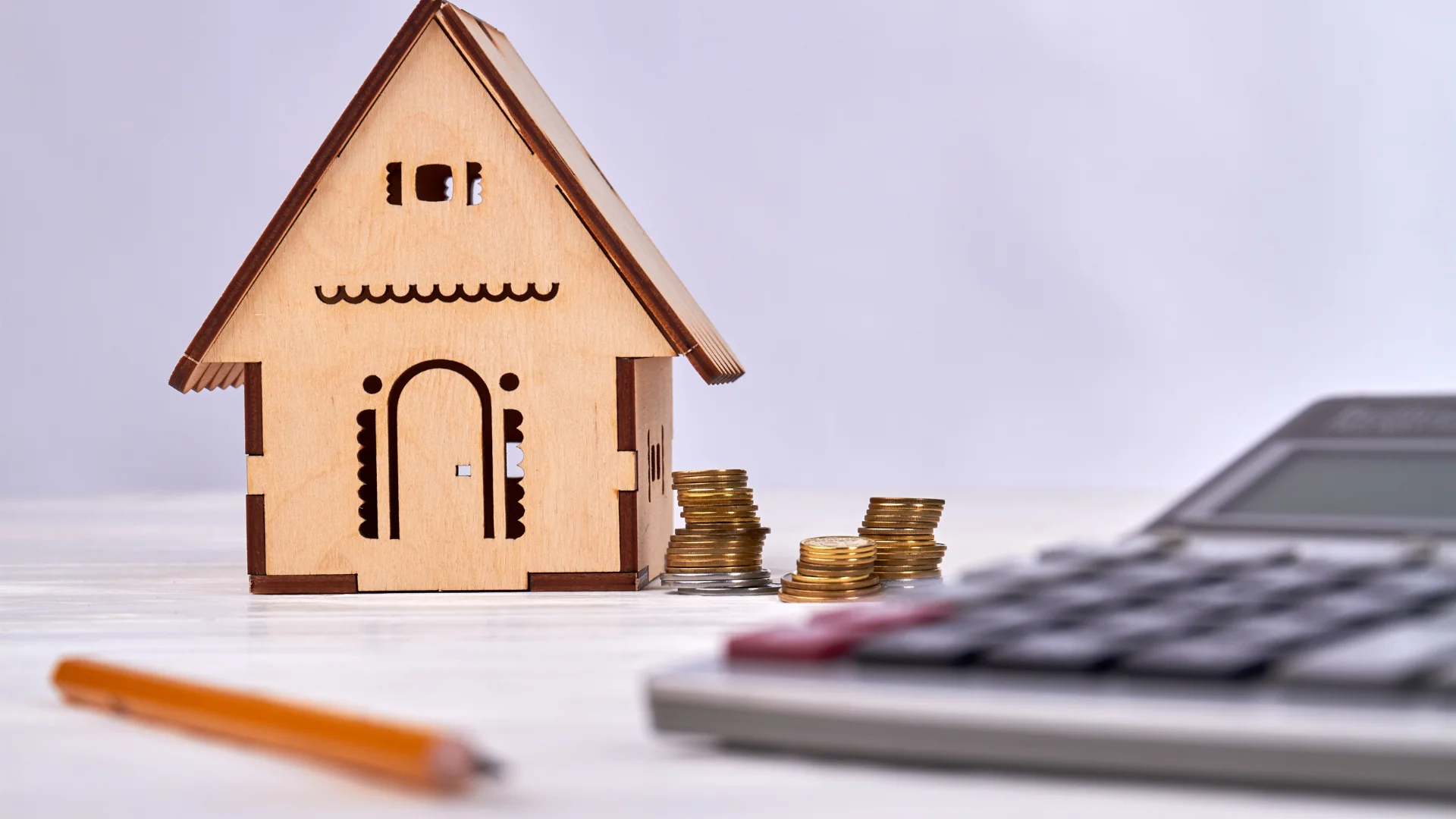 Kleines Holzhaus-Modell mit Münzstapeln daneben, im Vordergrund ein Taschenrechner und ein Stift – Symbol für Wertermittlung, Wert1914 und Versicherung.