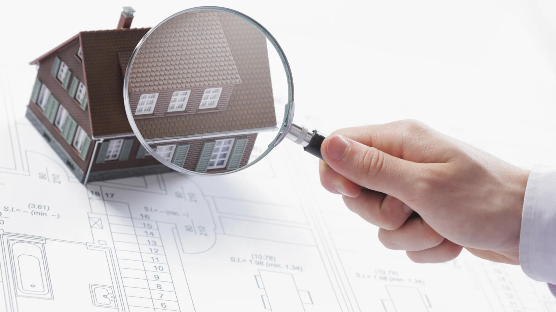 Hand hält eine Lupe über ein Miniaturhaus auf einem Bauplan – Symbol für Gebäudebewertung, Unterversicherung und Versicherungssummenprüfung.